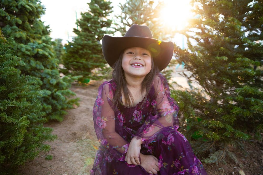 A joyful young girl in a purple floral dress and black cowboy hat smiles among green Christmas trees.