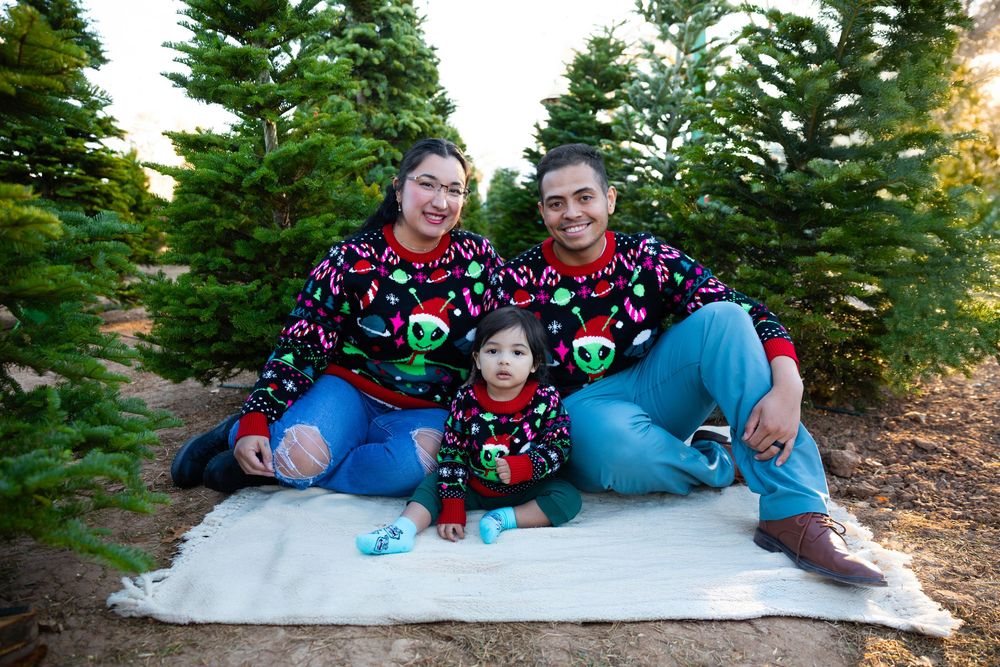 Family of three in matching Christmas sweaters sitting on a blanket among green Christmas trees at a tree farm.