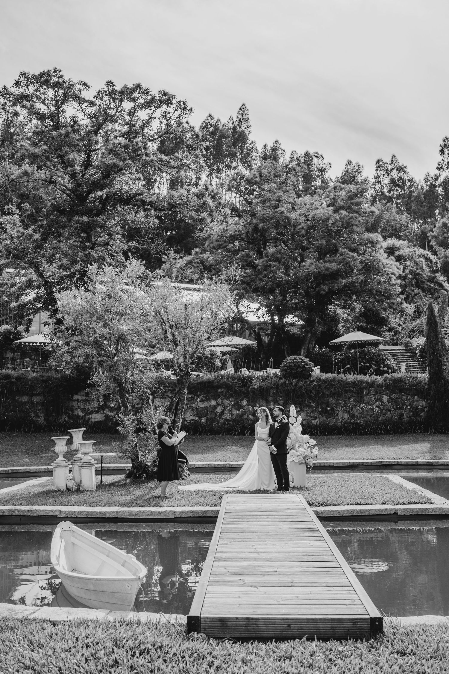 Elegant outdoor wedding cocktail hour on lush green lawn with white umbrellas, marquee tents, and guests mingling near a greenhouse.