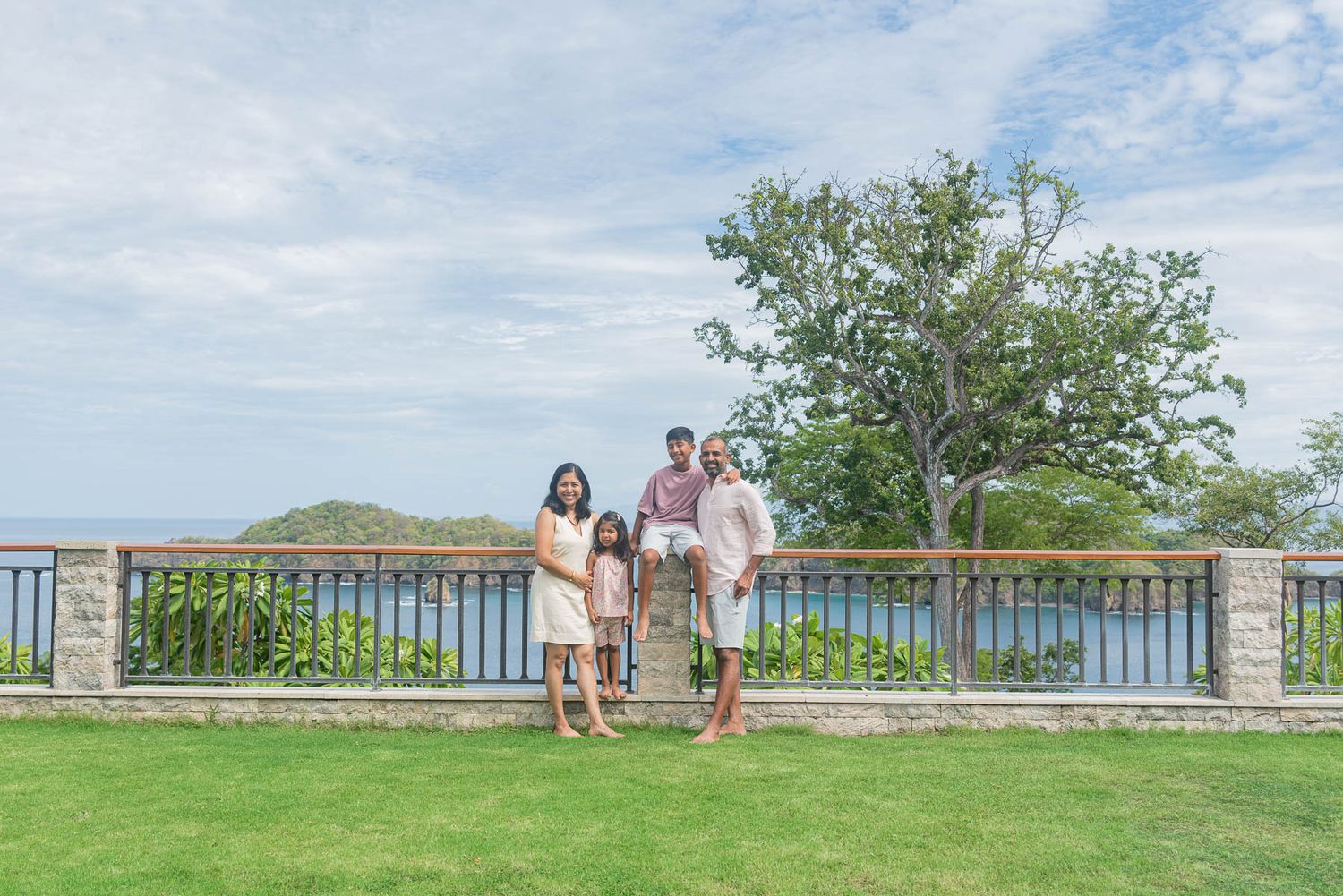 Family of four posing on a scenic overlook with lush greenery and ocean views under a partly cloudy sky.