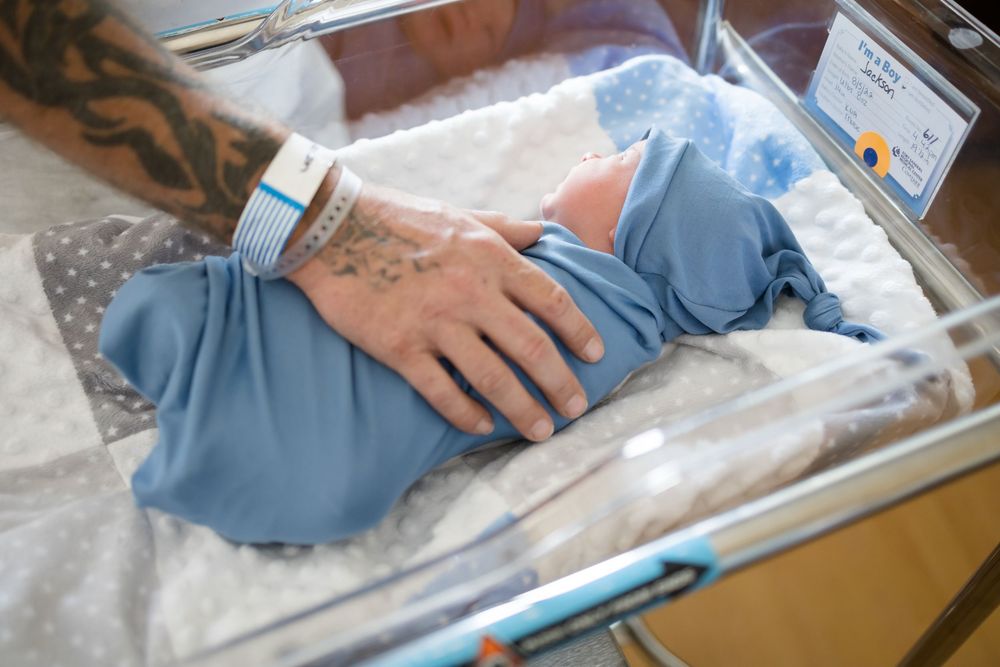 Tattooed parent's hand resting on swaddled newborn baby in hospital bassinet during a Fresh 48 Session at Fort Sanders in Knoxville, TN.