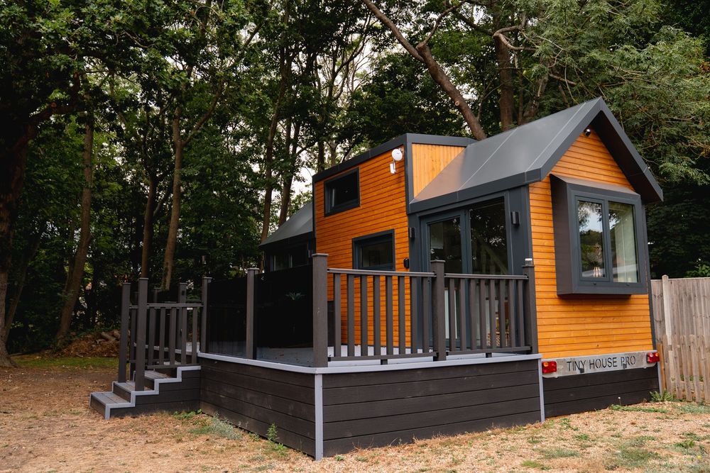 A modern tiny house with orange siding and dark trim, featuring a wraparound deck, nestled among tall trees.