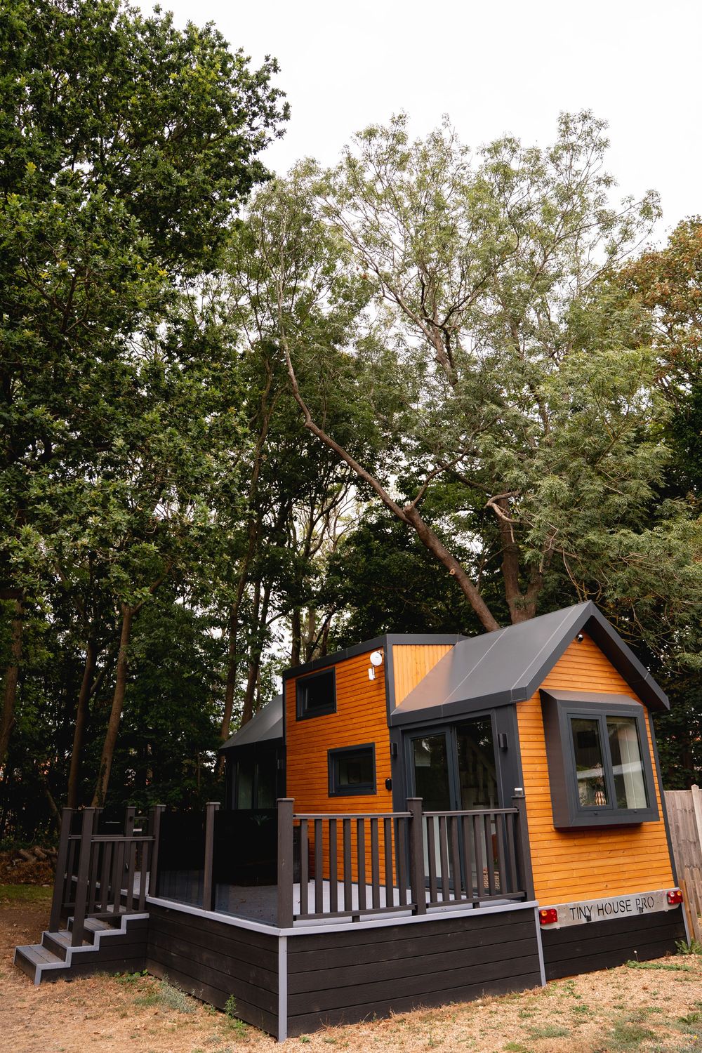 A charming orange tiny house with gray trim and deck, nestled among tall green trees in a wooded setting.