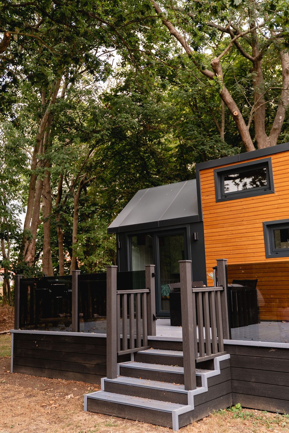 A modern tiny home with orange wood siding, dark deck, steps, and canopy entrance surrounded by tall green trees.