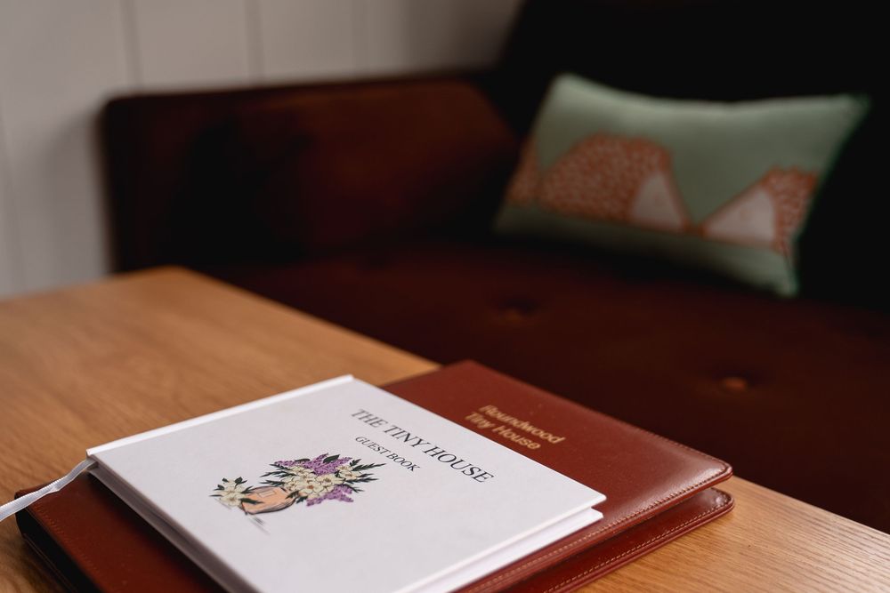 A tiny house guidebook rests on a wooden coffee table beside a brown leather journal, with a cozy sofa in the background.