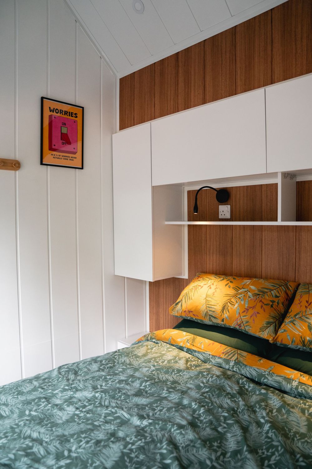 Cozy bedroom with green floral bedding, yellow pillows, built-in white shelving, wood panel headboard, and a pink framed wall poster.