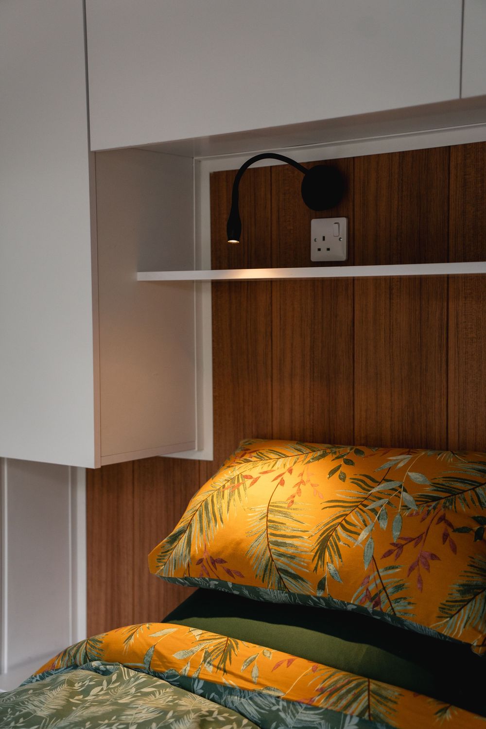 Cozy bedroom nook with wood panel headboard, tropical yellow floral pillows, wall-mounted reading lamp, and white shelving unit.