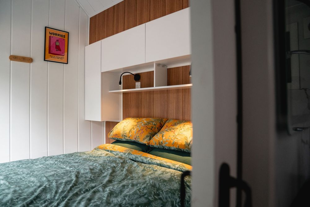 Cozy bedroom with green bedding, yellow floral pillows, built-in wooden shelving, and white cabinetry overhead.
