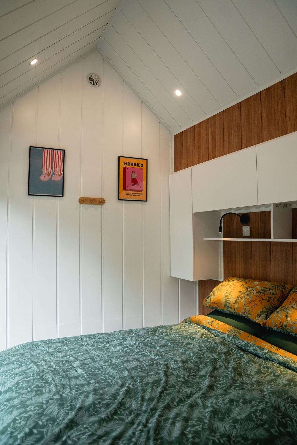 Cozy bedroom with green floral bedding, white shiplap walls, wood cabinetry, and colorful art prints under a vaulted ceiling.