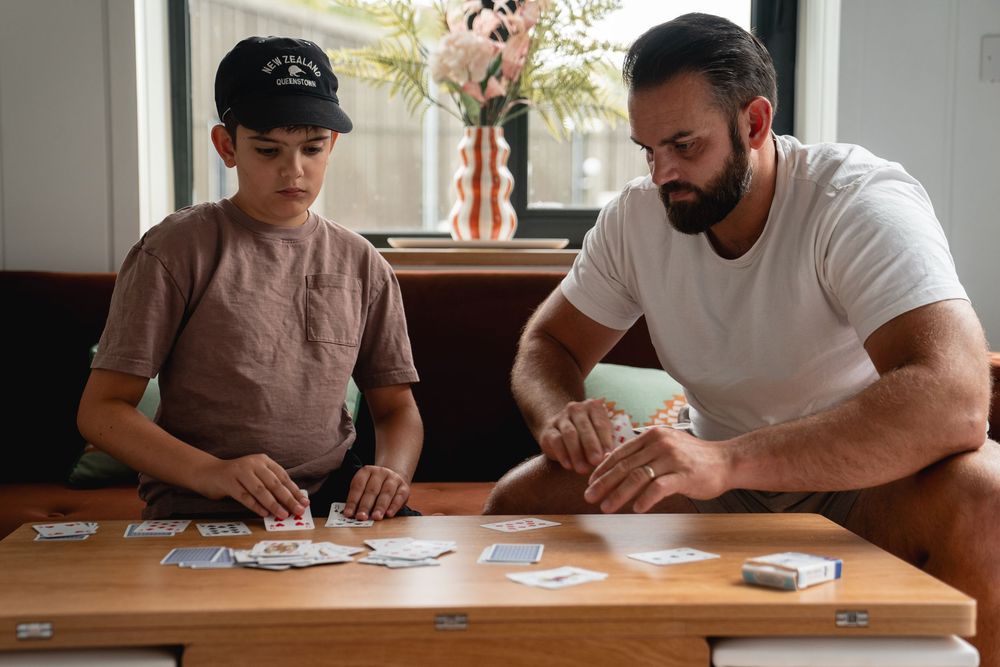 A man and boy play a card game together at a wooden table indoors.