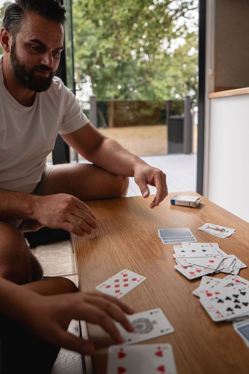 Bearded man in white t-shirt playing cards at wooden table with another person, green trees visible in background.