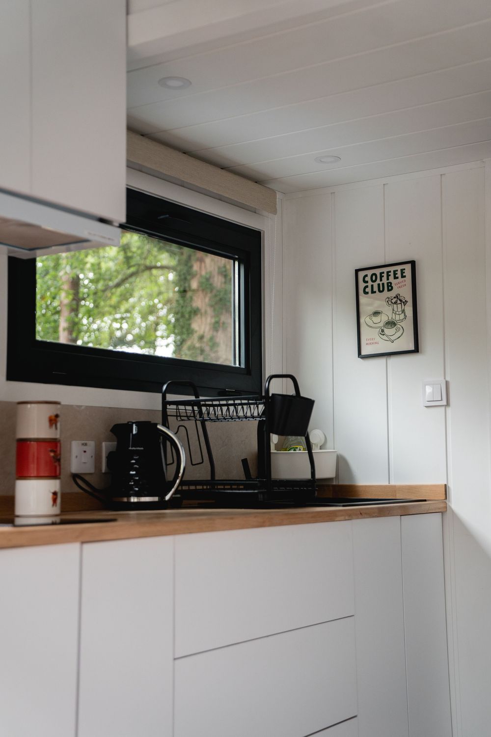 Modern white kitchen corner with wood countertop, black kettle, dish rack, and Coffee Club wall art near a window.