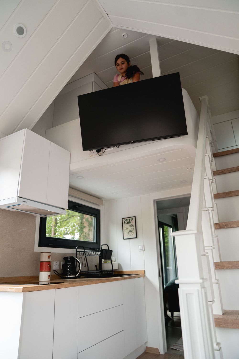 Tiny house interior with loft bedroom, white kitchen cabinets, wall-mounted TV, and woman looking down from upper level.