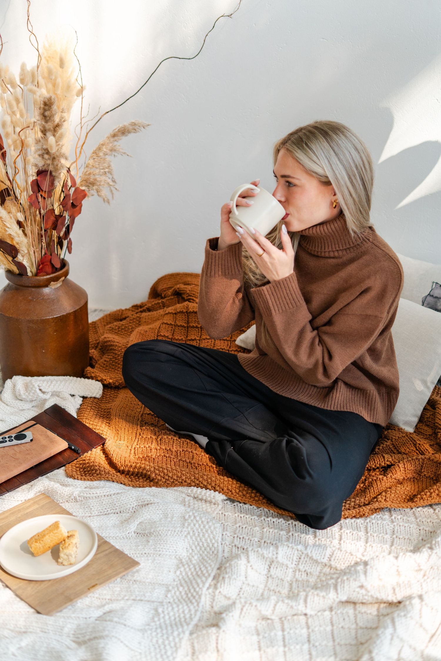 Woman in brown turtleneck sweater sipping from white mug while sitting cross-legged on cozy throws with autumn decor.