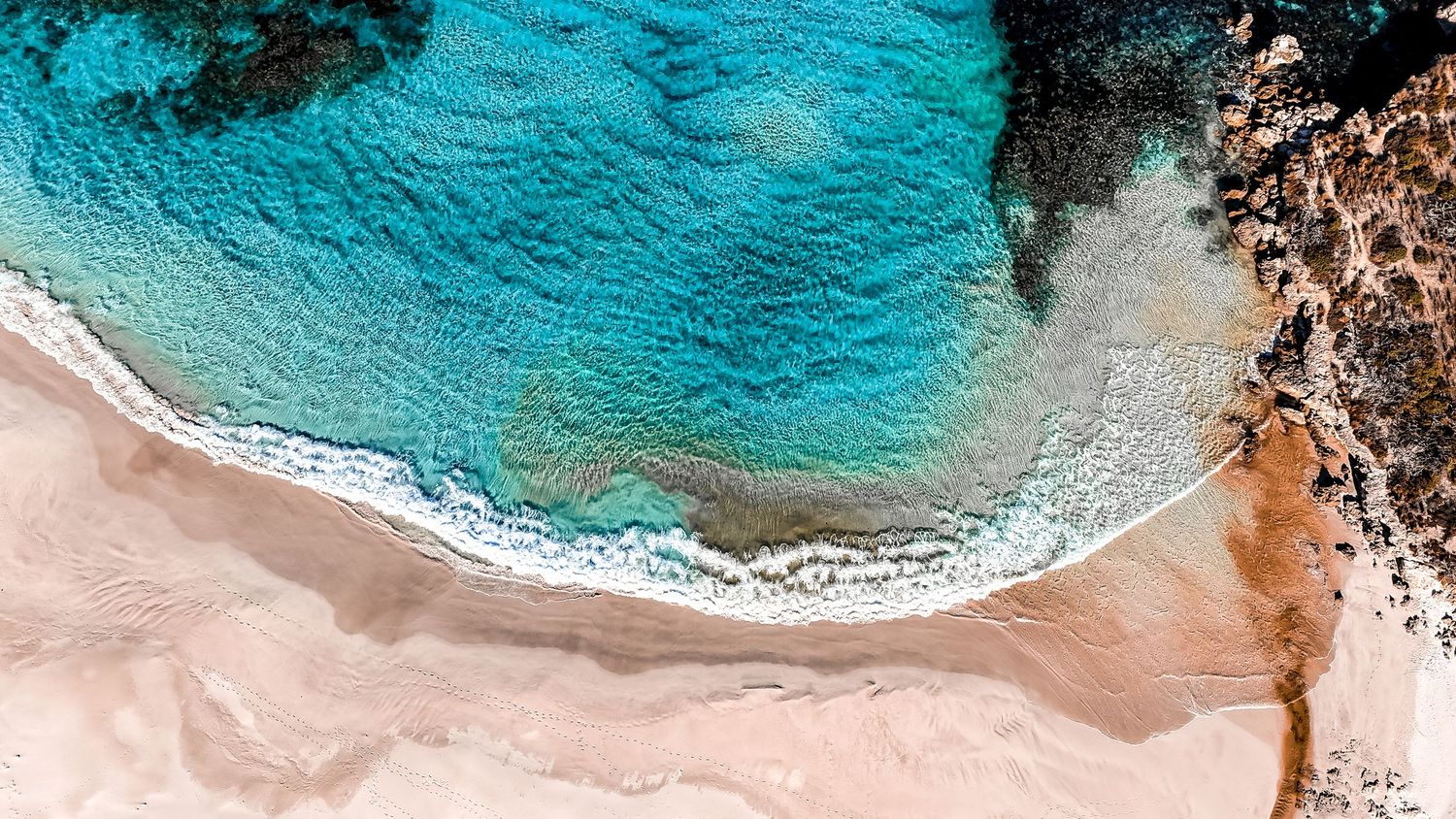 Aerial view of turquoise ocean waves meeting a sandy beach shoreline with rocky outcrops.