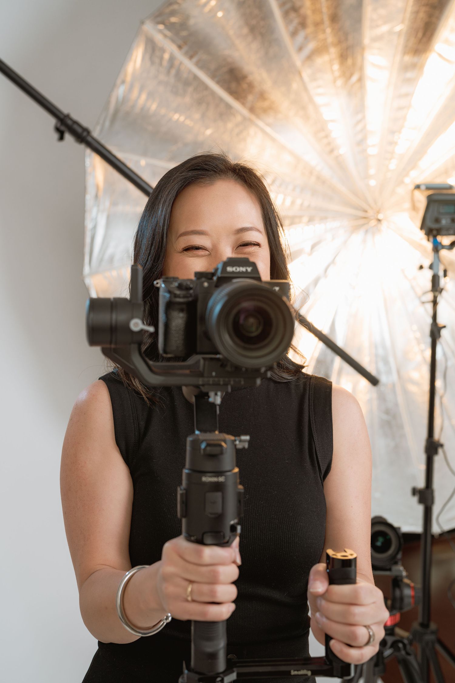 Woman smiling behind a Sony camera on a gimbal stabilizer in a photography studio with reflective lighting equipment.