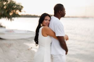 Couple standing close together on a white sand beach at sunset near a small boat.