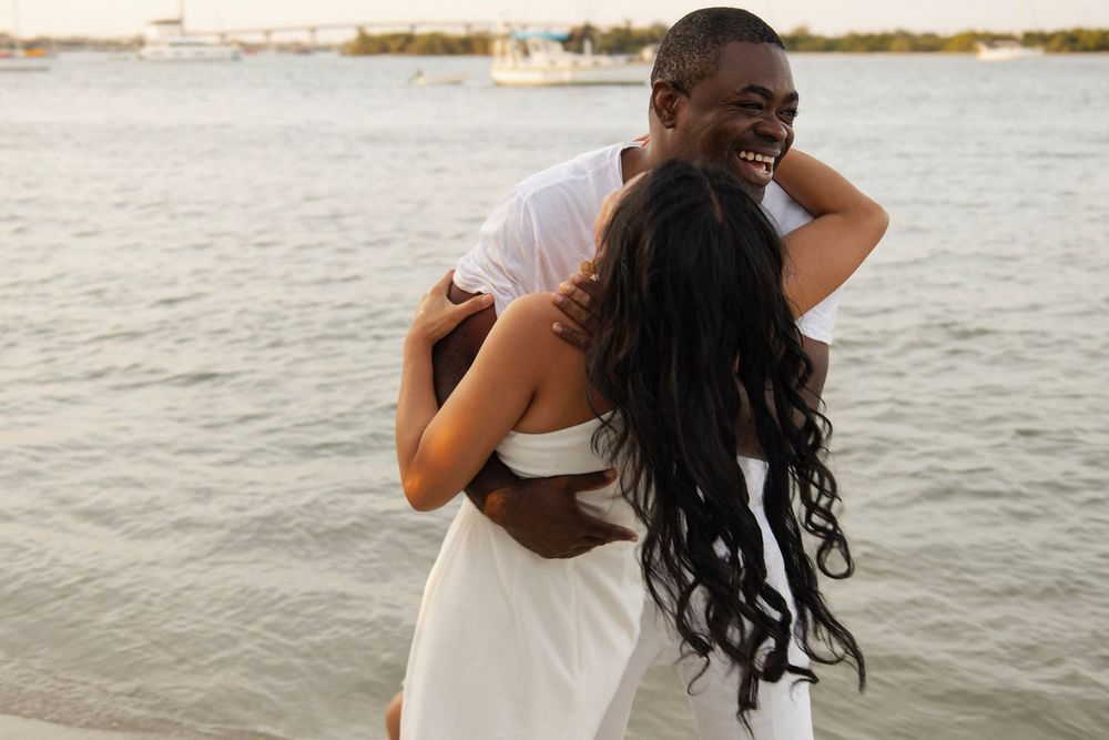 Happy couple hugging and spinning joyfully on a beach with calm water in the background.