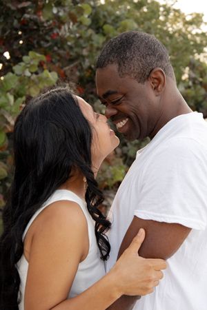 Smiling couple holding each other close and laughing together outdoors near colorful foliage.