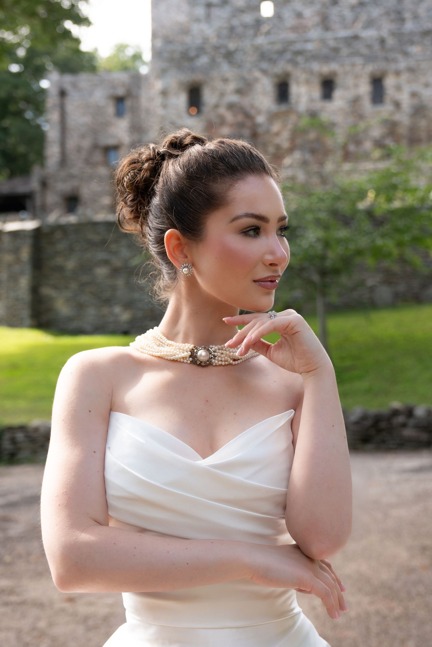 Elegant bride in strapless white gown with pearl necklace poses gracefully before a stone castle wall.