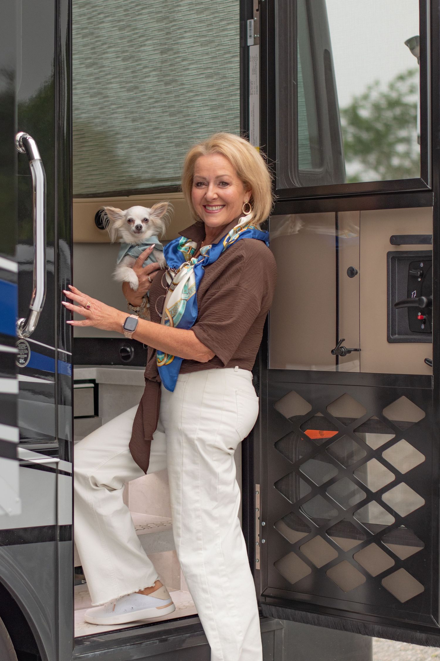 Smiling blonde woman holding a small dog while stepping out of a luxury RV, wearing white pants and a colorful scarf.