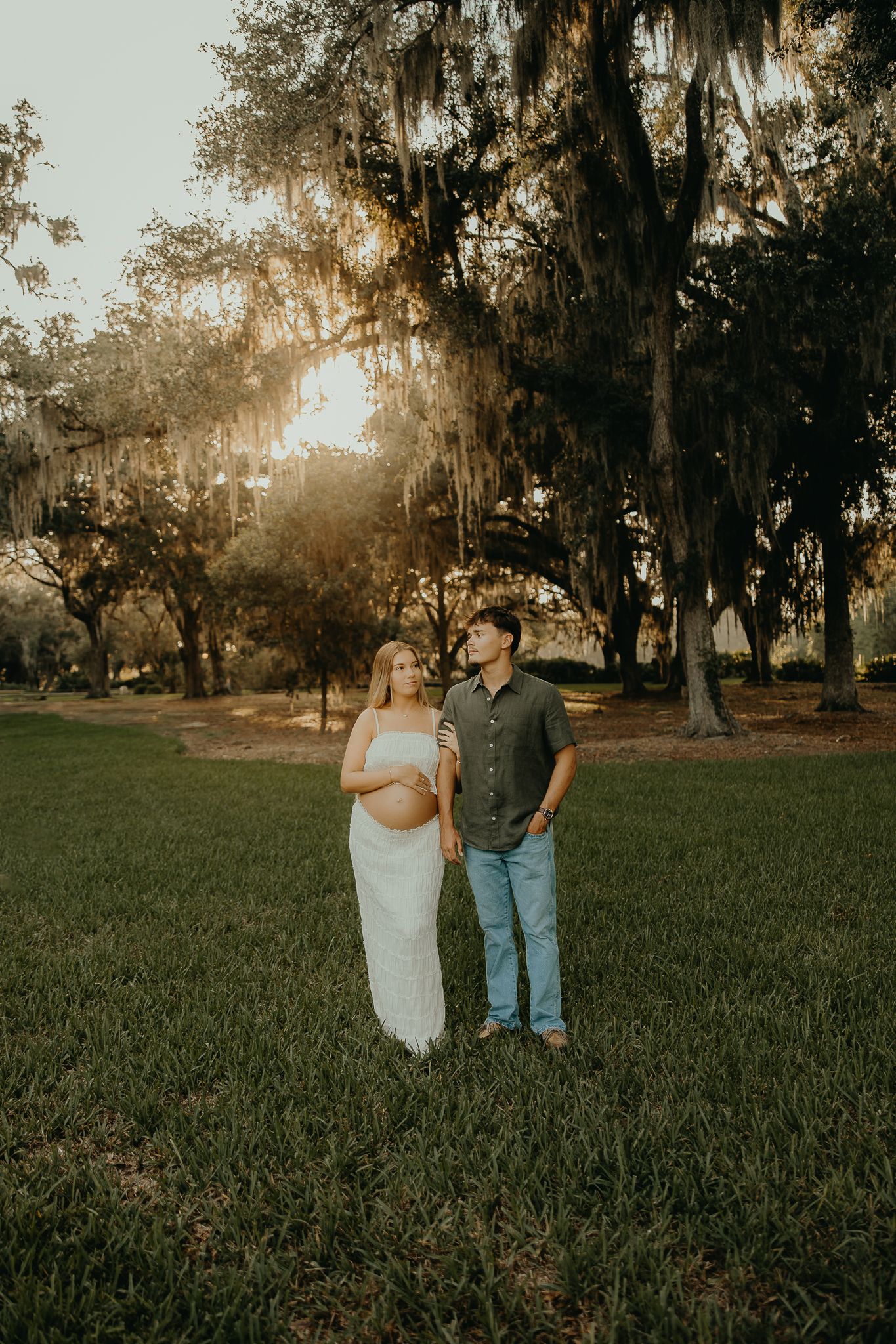 Pregnant couple stands in sunlit park with Spanish moss-draped oak trees during golden hour maternity photo session.