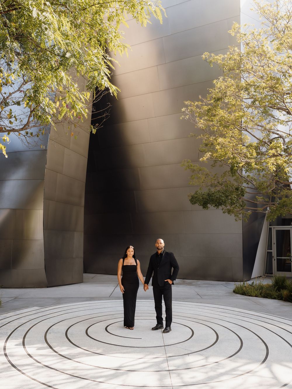 A couple in black attire holds hands on a circular patio outside the Walt Disney Concert Hall in Los Angeles.
