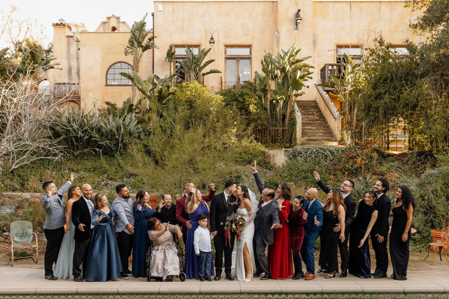 Large wedding party poses outdoors at elegant venue with lush gardens and Spanish-style architecture during golden hour.