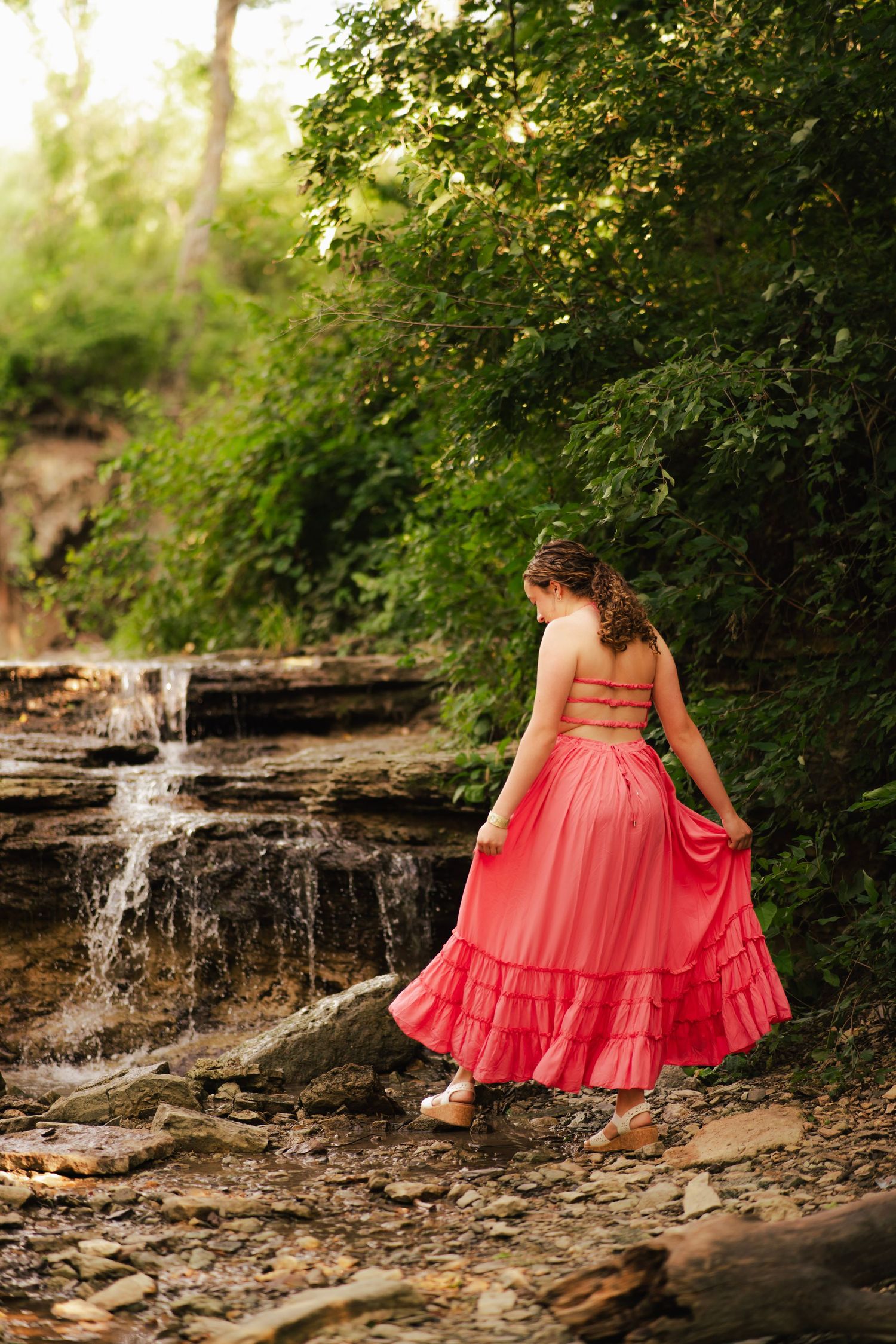 Woman in pink ruffled maxi skirt and crop top standing on rocks near a small waterfall surrounded by lush greenery.