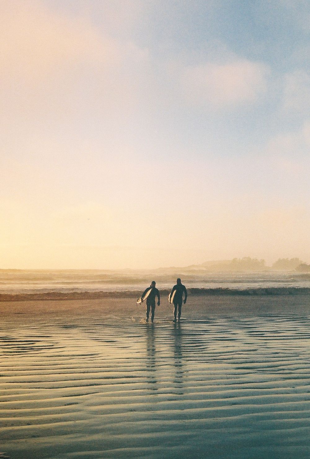 Two surfers carrying boards walk along a misty beach at golden sunset, silhouetted against the glowing horizon.