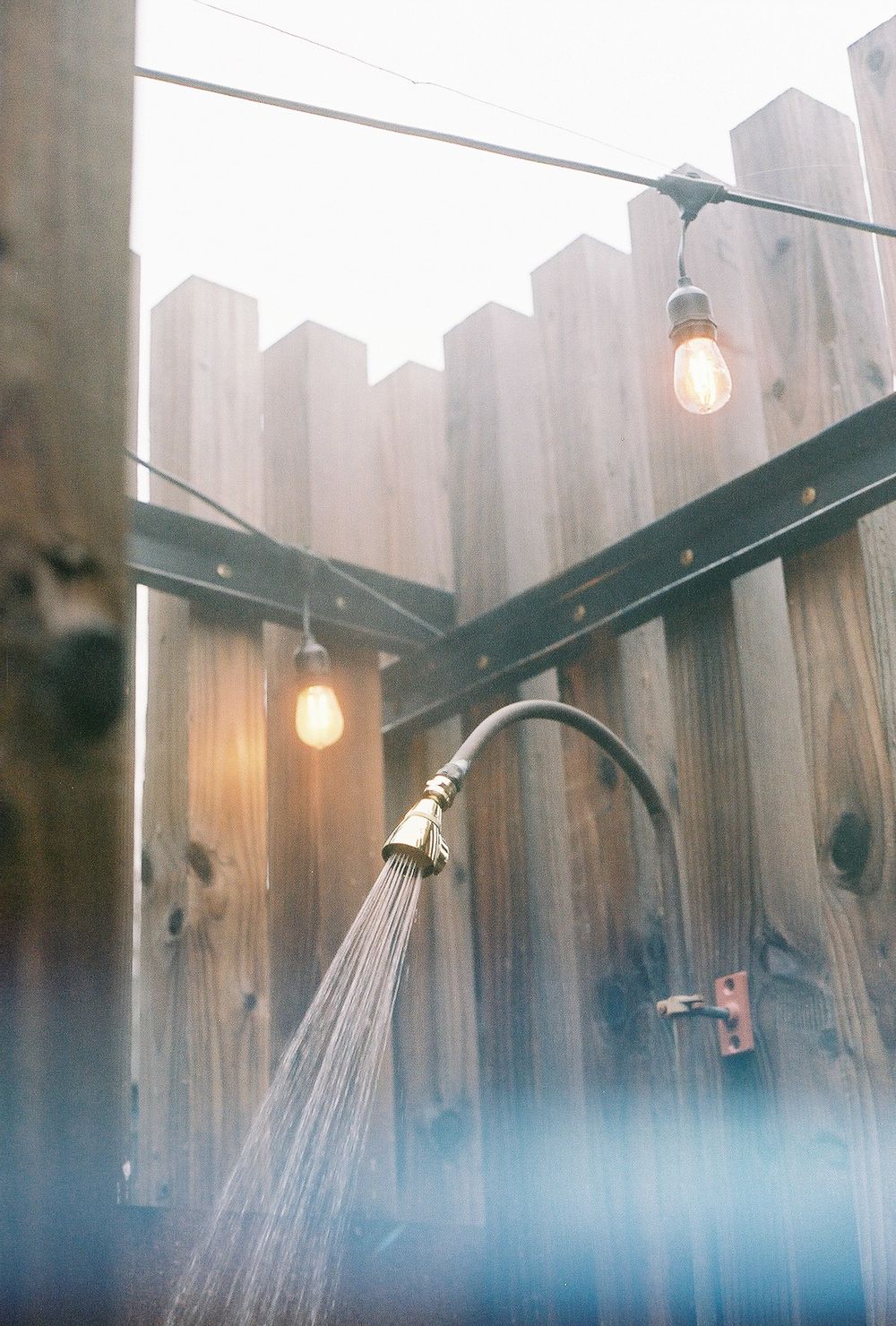 Outdoor shower with brass showerhead spraying water, Edison bulb string lights, and rustic wooden fence backdrop.