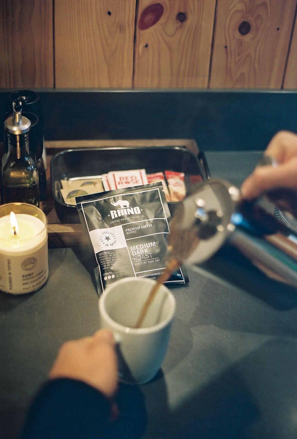 Hands pouring hot water into a white mug beside Rhino Medium Dark coffee bag and lit candle on counter.