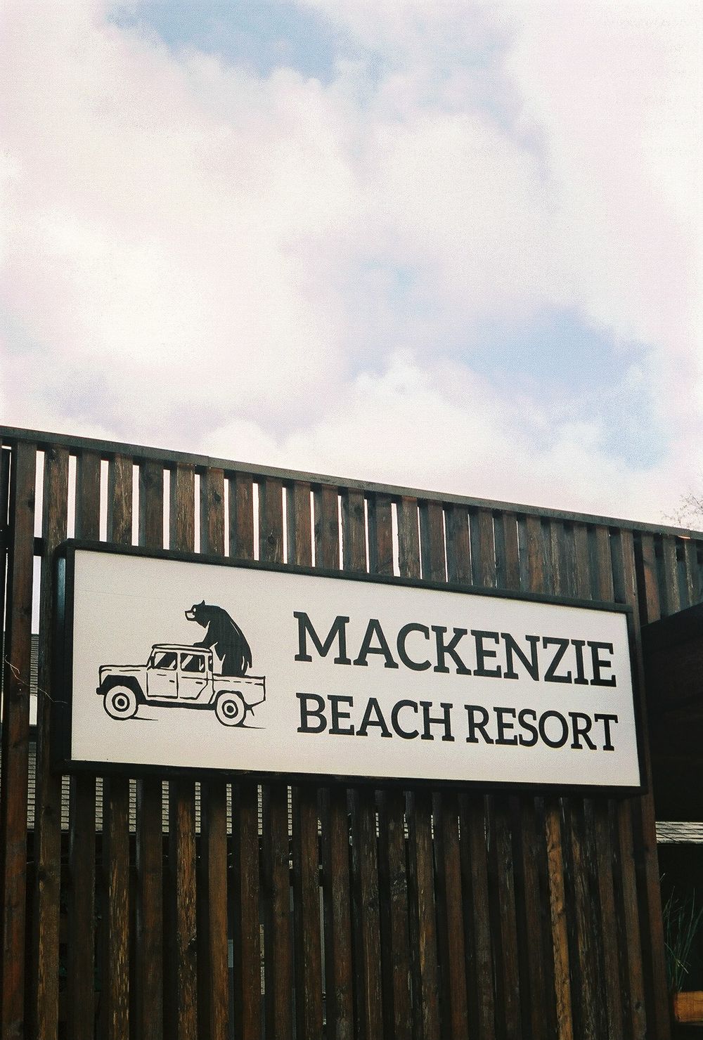Sign for Mackenzie Beach Resort on a wooden fence with a bear and jeep logo against a cloudy sky.
