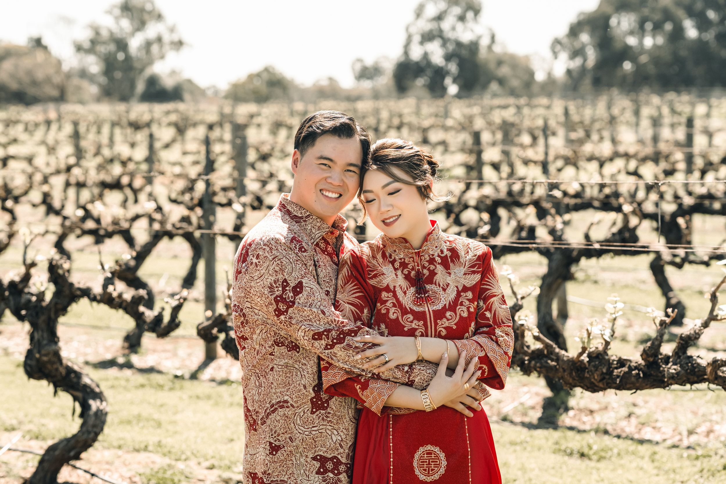 A couple in traditional red Chinese wedding attire embraces in a vineyard, smiling warmly on a sunny day.