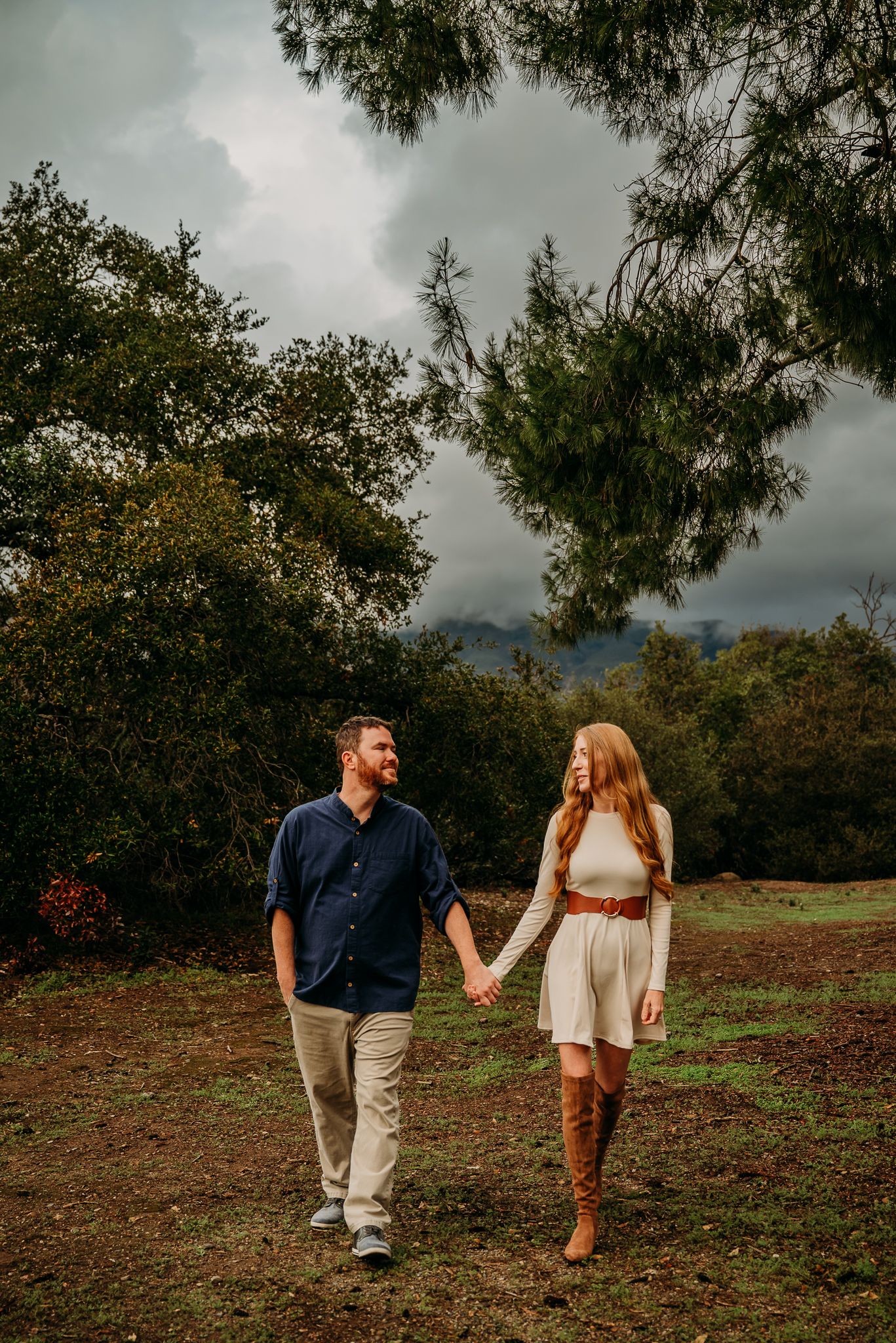 Couple holding hands walking through a lush green park with mountains and dramatic cloudy sky in the background.