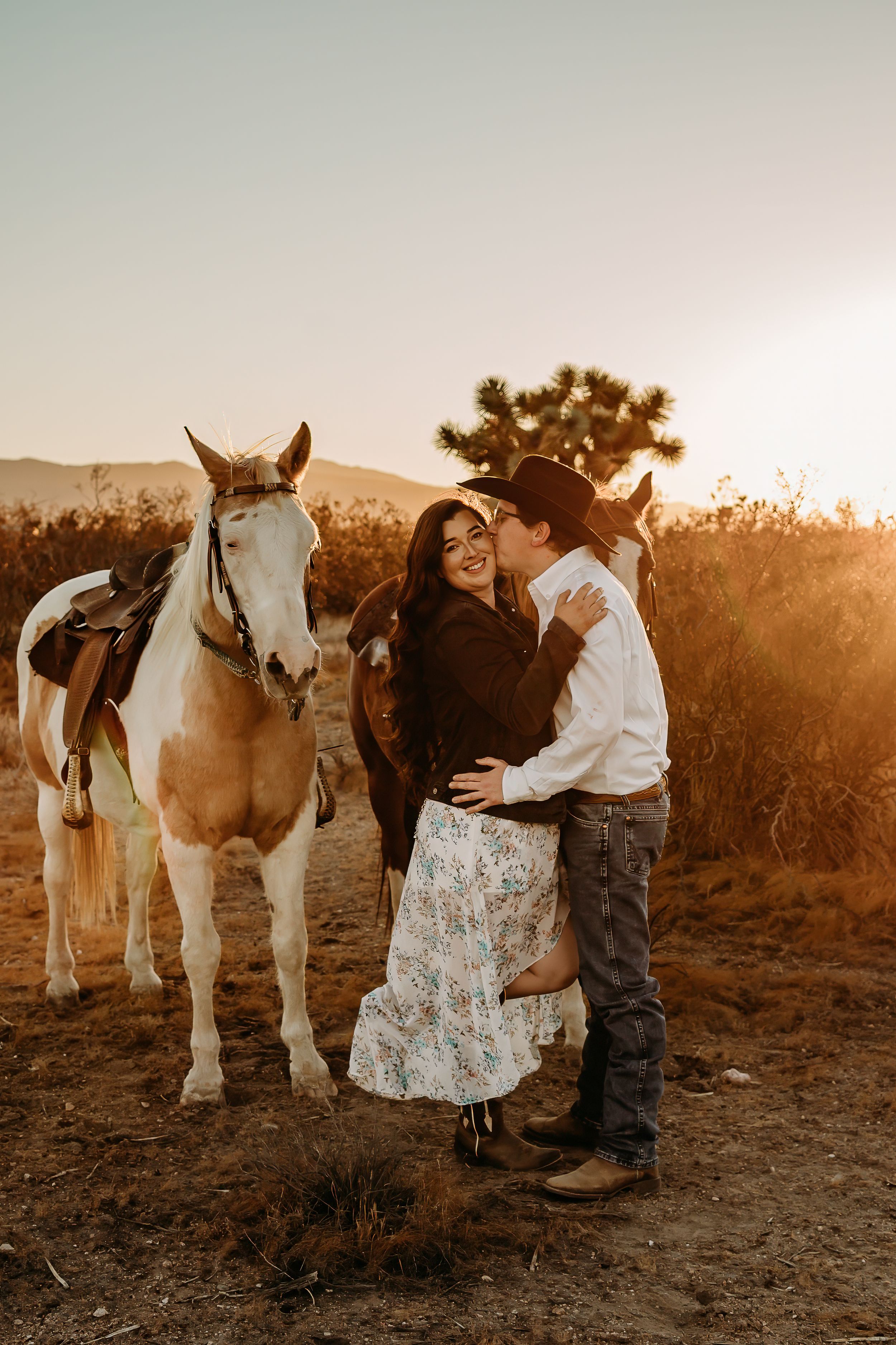 Couple in western attire sharing a romantic moment at sunset beside two horses in a golden desert landscape.