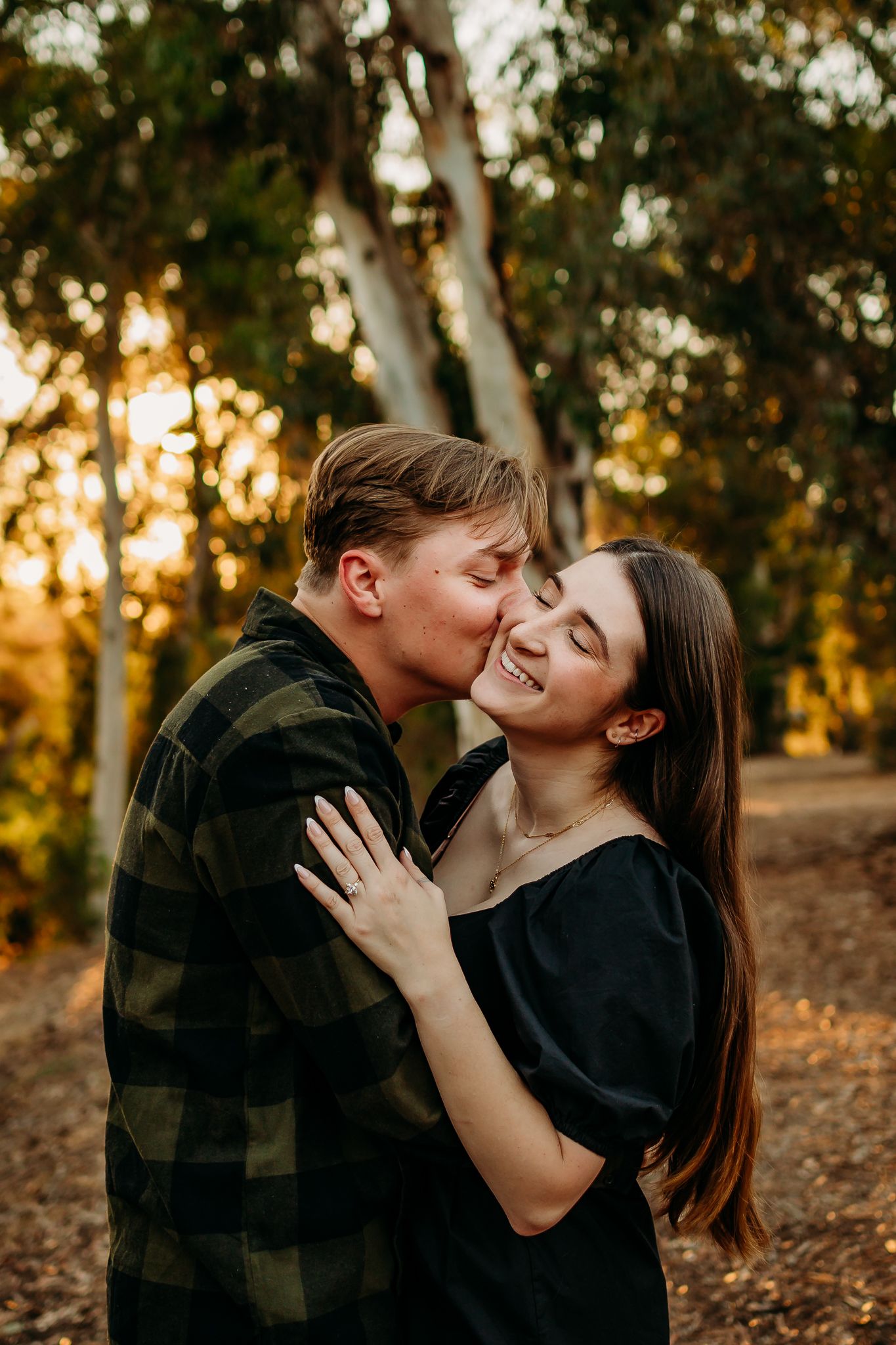 Couple sharing a romantic kiss at golden hour in a forest, woman smiling joyfully in black top.