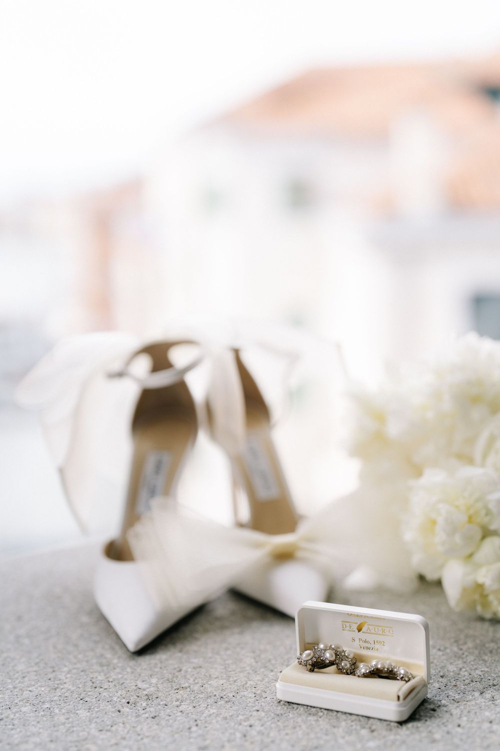 White bridal shoes and delicate jewelry box arranged with fresh white flowers on concrete surface.