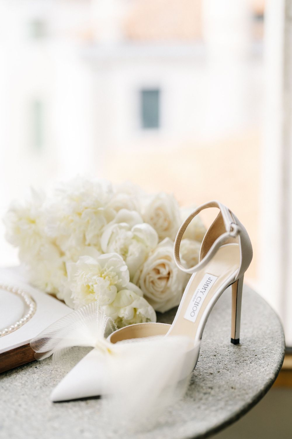 Elegant white Jimmy Choo bridal heel displayed next to white peonies on a gray surface.