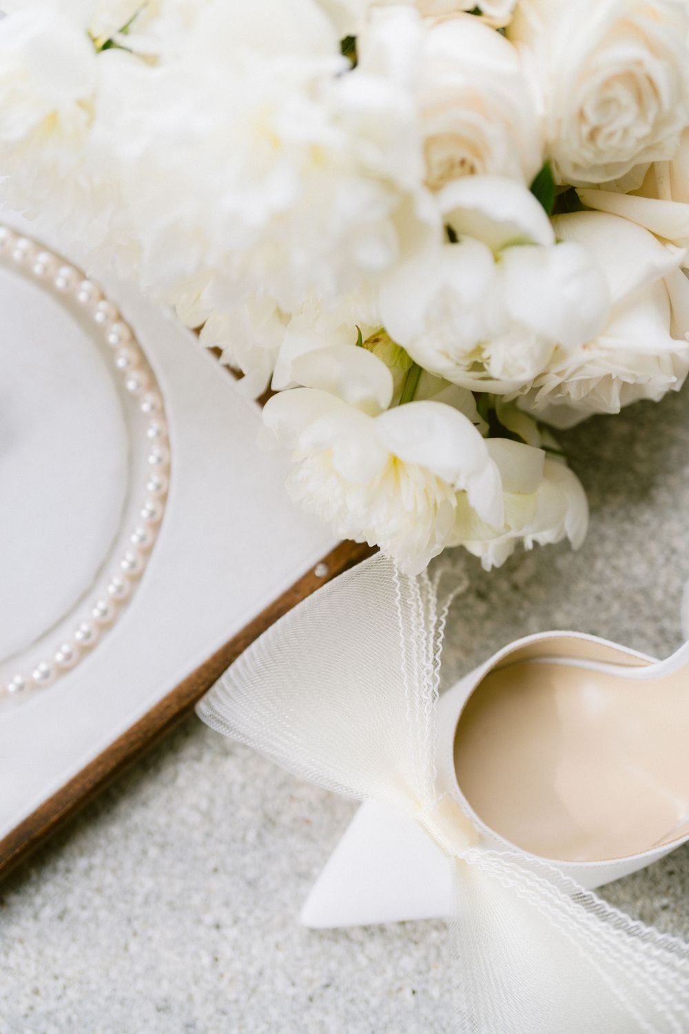 White roses and pearl-trimmed plate arranged with elegant bridal shoe on textured gray surface. Photoshoot by Venice Photographer Alina Indi