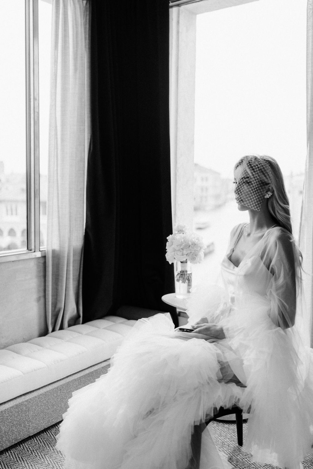 Black and white portrait of person in wedding dress sitting by window with bouquet. Photoshoot by Venice Photographer Alina Indi