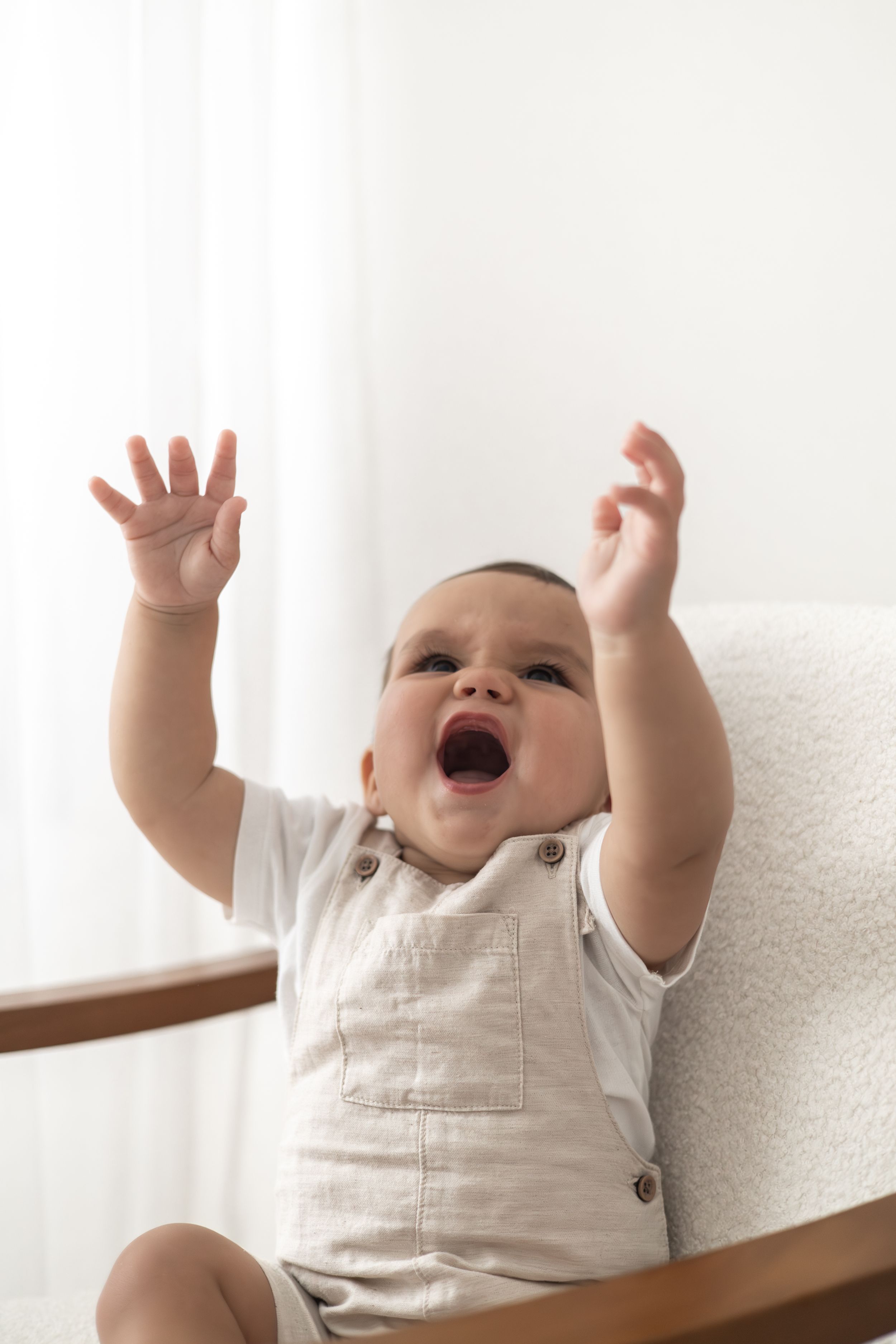 A joyful baby in beige dungaree sits on a white chair, arms raised and mouth open in excitement first birthday photography Nottingham