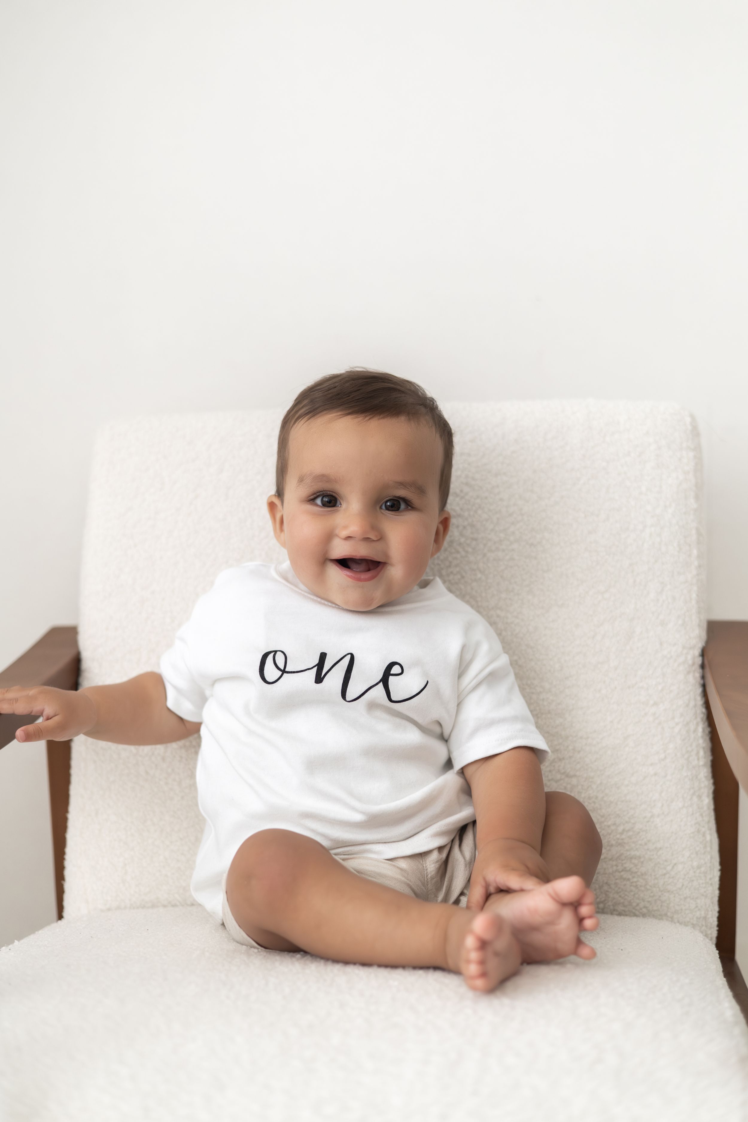Smiling baby wearing a white 'one' T-shirt sitting on a cream chair celebrating their first birthday first birthday photography Nottingham
