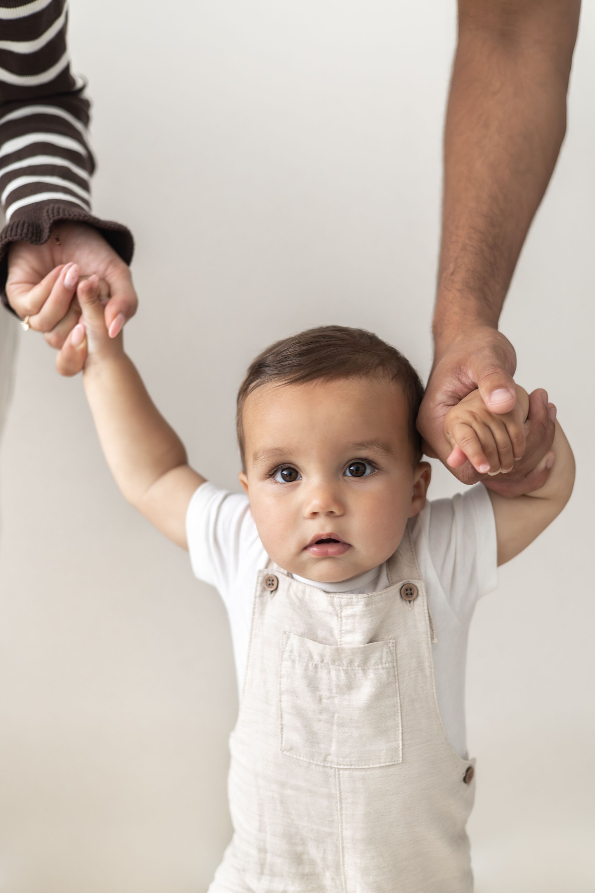 Adorable baby in white dungarees holding parents hands against white background first birthday photography Nottingham