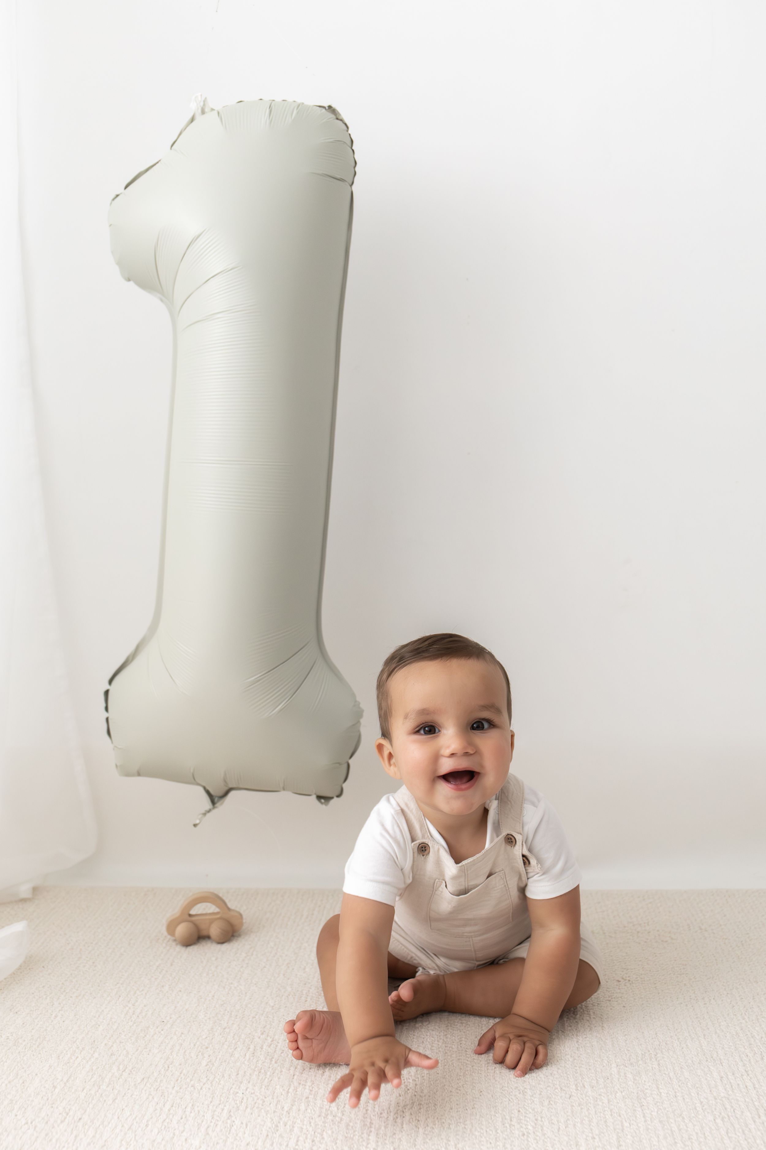 Smiling baby sitting on white floor next to a number one balloon celebrating their first birthday first birthday photography Nottingham