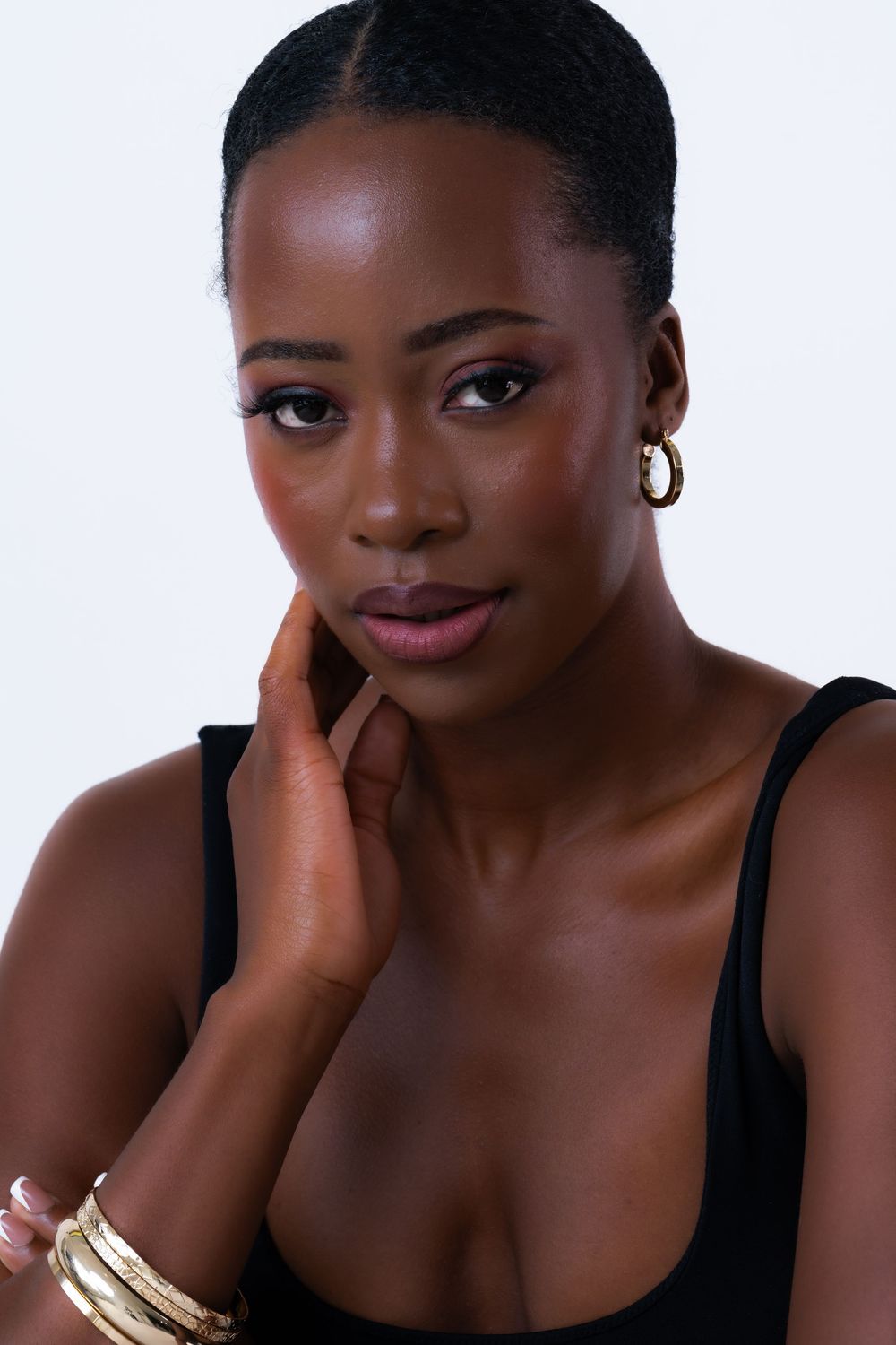Young Black woman in black tank top with gold hoop earrings and bangles, hand on cheek, posing against white background.