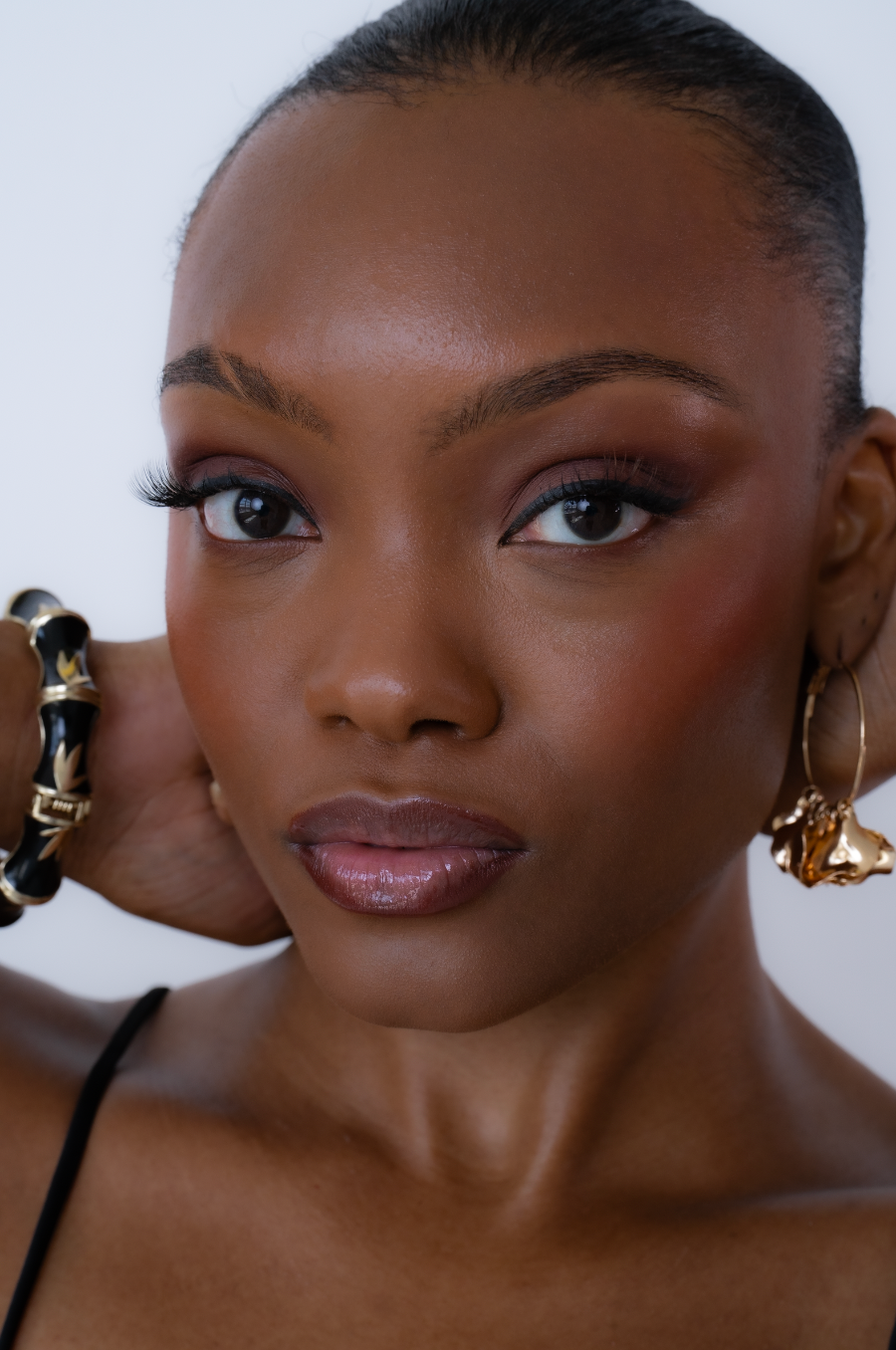 Close-up portrait of a young woman wearing gold hoop earrings and a black and gold bracelet with flawless makeup.