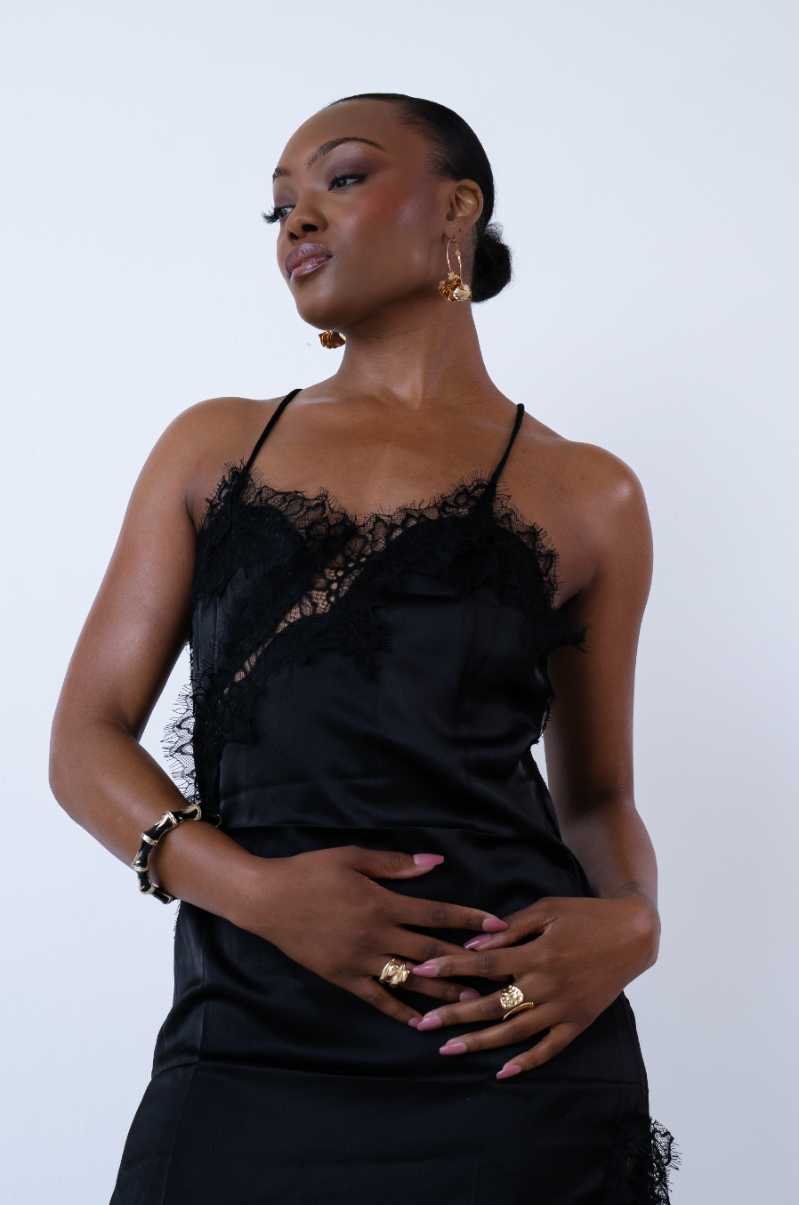 Woman in black lace satin slip dress with gold jewelry posing against white background.