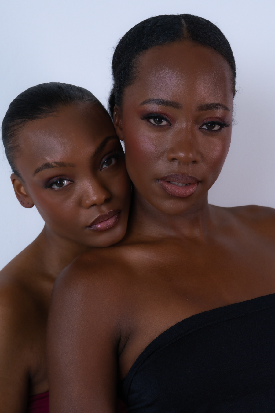 Two Black women with sleek updo hairstyles and flawless makeup pose together against a white background.