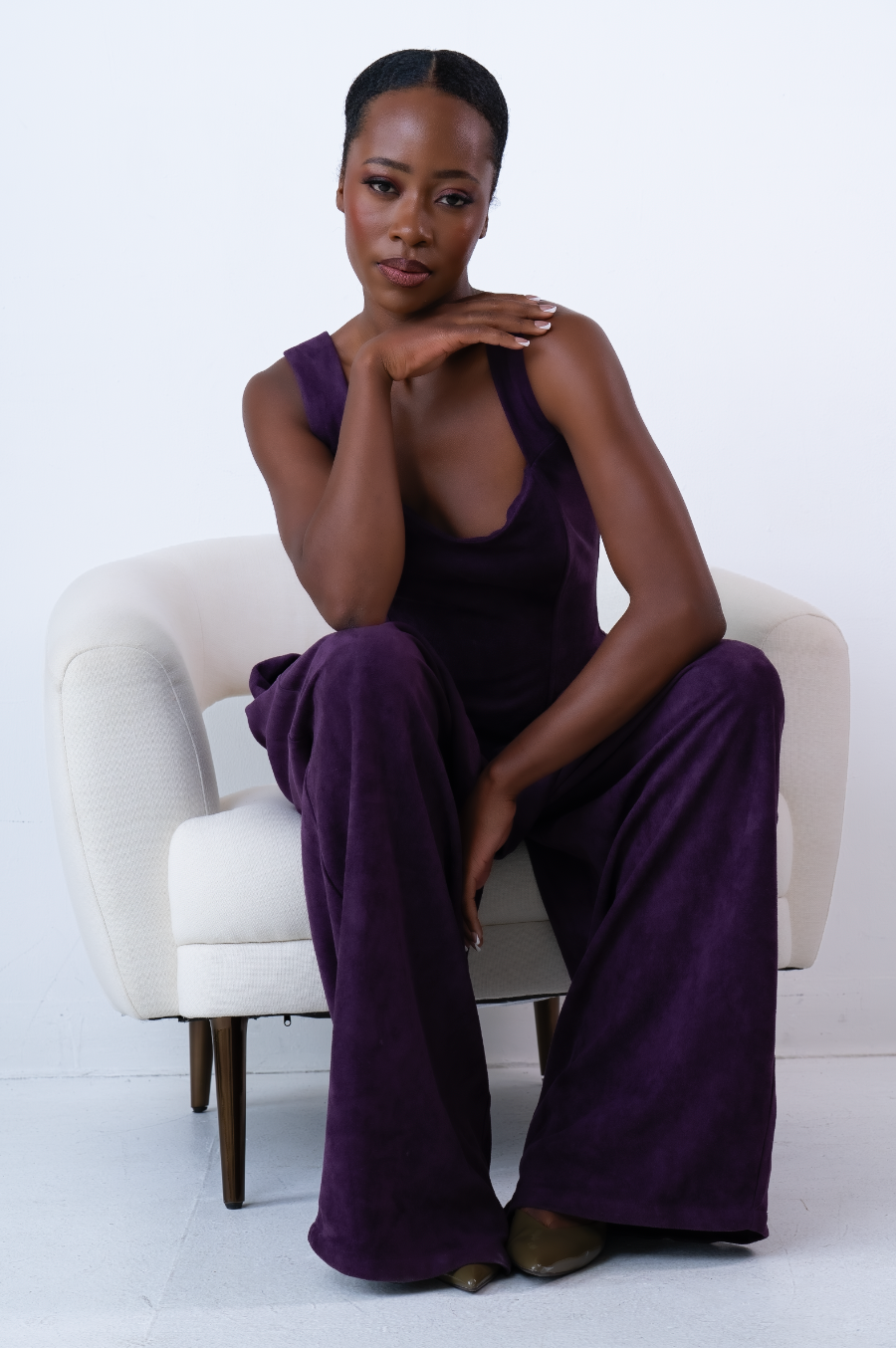 Woman in purple velvet jumpsuit seated gracefully in white chair, posing with hand under chin.